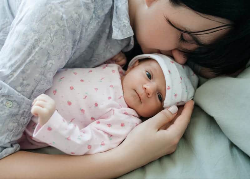 cluster feeding baby in pink onesie with strawberries and mother hugging her lying on bed