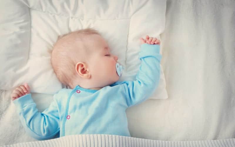 cluster feeding baby lying on bed with a pacifier and arm up in teal onesie